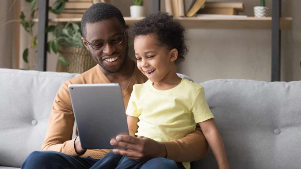 Daughter sits in mans lap on gray couch while they look at silver tablet | User Activation for Streaming Companies