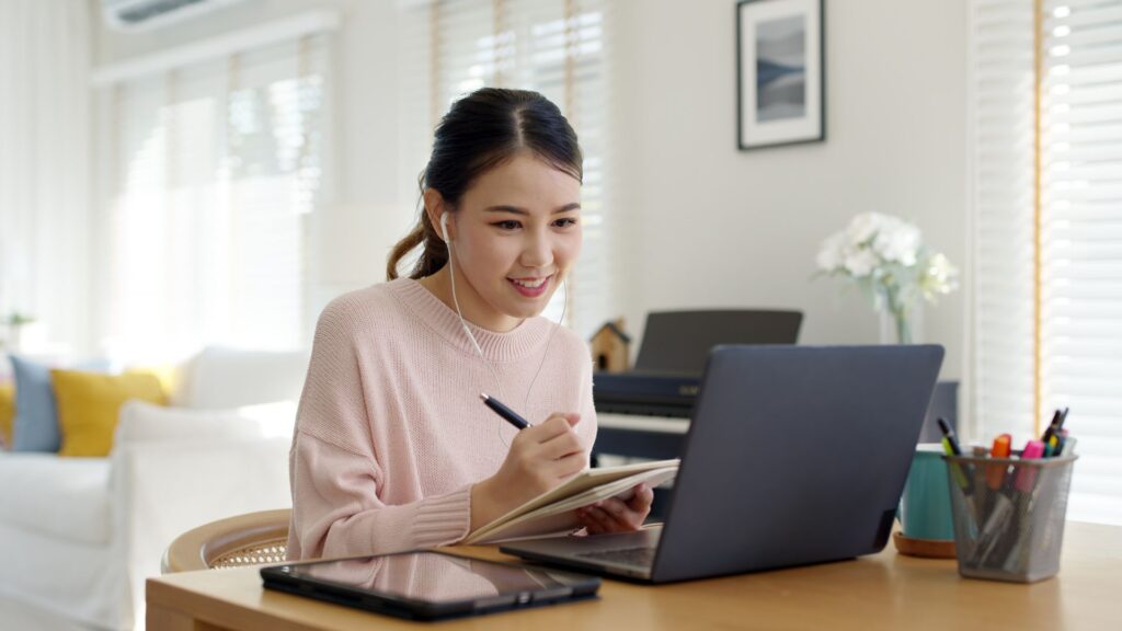 Woman wearing headphones smiles while taking notes and using laptop | User Resurrection for SaaS Companies