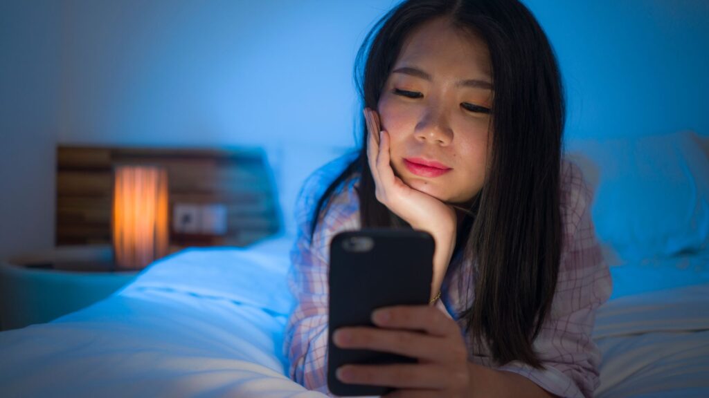 Girl’s face is backlit as she looks at smartphone while laying on bed in cozy, dark room | Finding the Right SaaS Users